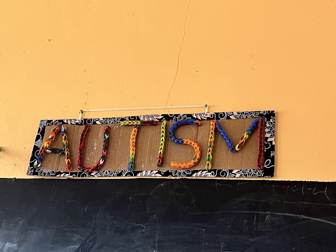 The word "AUTISM" spelled out in colorful plastic band weavings set against a cardboard rectangle background.
