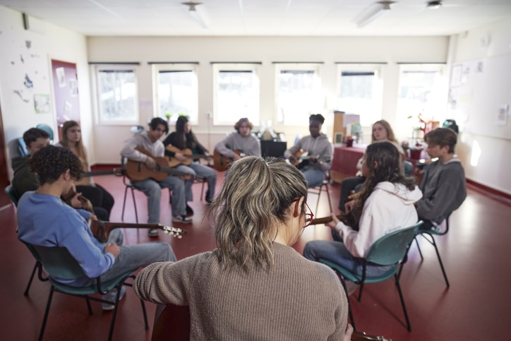 Teenagers sitting in chairs placed in a circle holding guitars in a bright music classroom